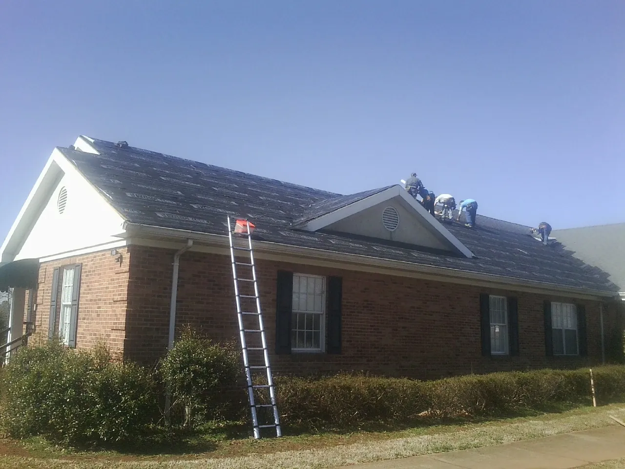 Crew tearing shingles off a brick ranch roof with a ladder set — Emergency Roof Repair work by our crew in Mount Solon