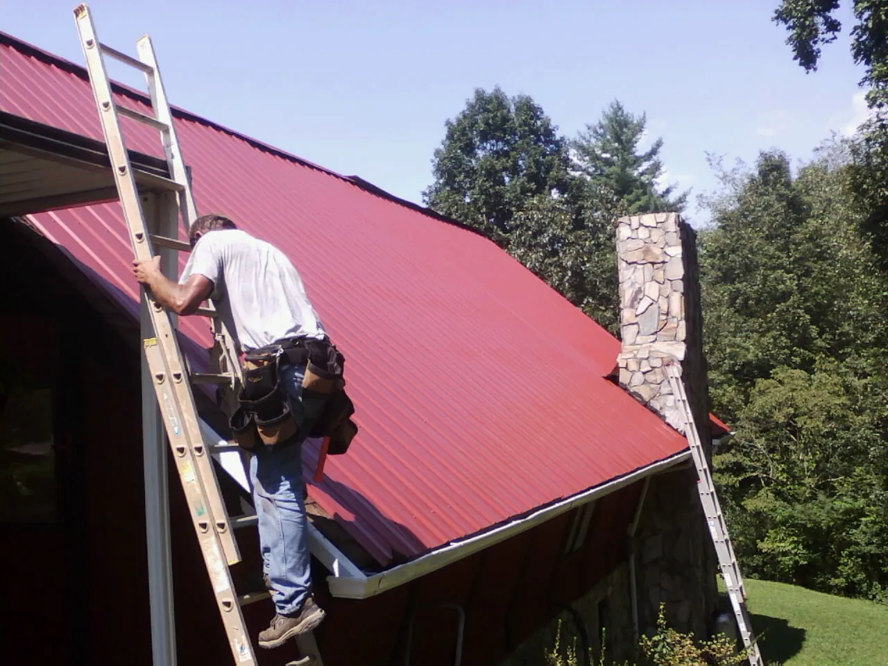Roofer on a ladder beside a red metal roof and stone chimney — Soffit & Fascia Repair by Roofing Brothers in Mount Solon